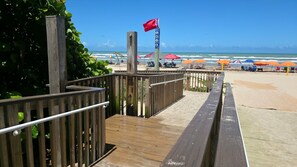 Ein Holzsteg führt zu einem Strand mit Sonnenschirmen und einer Flagge.
