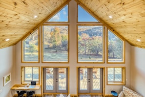 A high-ceilinged room with large windows, a wooden desk, and a view of a deck and trees. - BaseCamp Bryson-Panoramic Views-Pet Friendly-Paved-Hot Tub-Fire-pit (Bryson City)