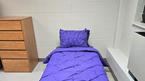 A single bed with a purple bedspread and matching pillow in a room with white brick walls and a wooden dresser.