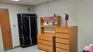 A dormitory room with a bed, a wooden dresser, a black closet, and a shelf with books and a vase.