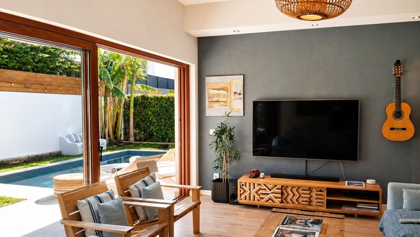 Ein modernes Wohnzimmer mit großem Fernseher, Holzmöbeln, einer Gitarre und Blick auf einen Pool.