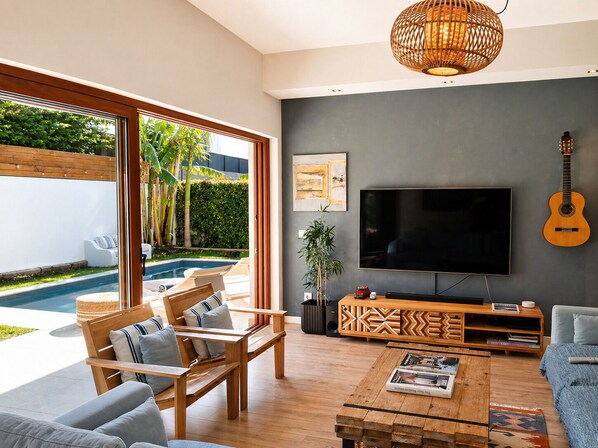 Un salón moderno con un televisor grande, muebles de madera, una guitarra y vistas a una piscina.