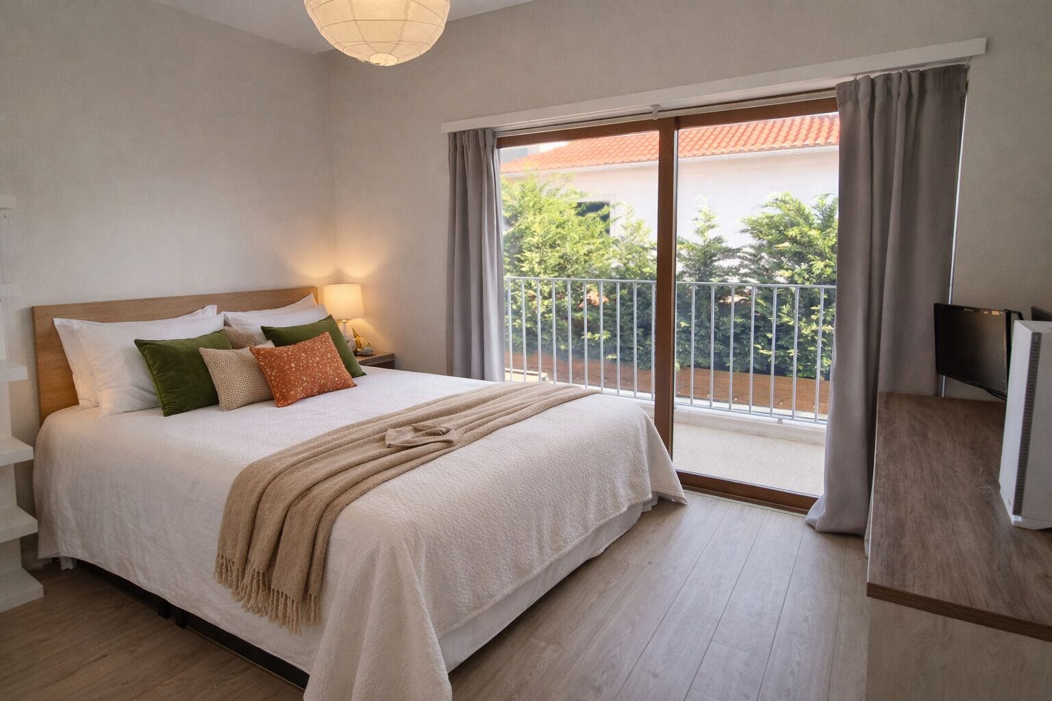 Un dormitorio con cama, almohadas, mesita de noche con lámpara, televisor y balcón con barandilla.