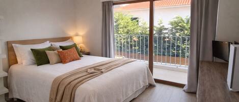 Un dormitorio con cama, almohadas, mesita de noche con lámpara, televisor y balcón con barandilla.