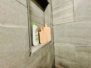 A bathroom with a tiled wall and a shelf holding toiletries.