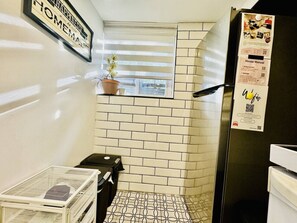 A compact kitchen with white tiled walls, a black refrigerator, a potted plant on a shelf, and a small window with blinds.