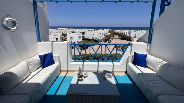 A balcony with white sofas and blue cushions, a small table with a model ship, and a view of the sea and buildings. - Spacious 3BR Apartment w Rooftop Terrace&Jacuzzi (Mersa Matruh)