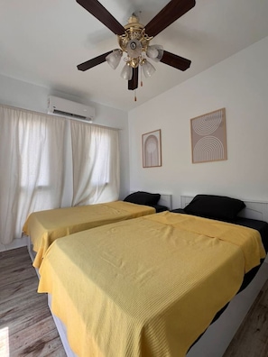 Une chambre avec deux lits, un ventilateur de plafond, un appareil de climatisation et deux œuvres d’art encadrées accrochées au mur.