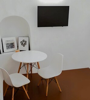 Een kamer met een witte tafel en twee witte stoelen, een zwarte tv aan de muur bevestigd en twee ingelijste foto's.