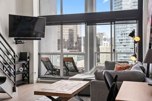 A modern living room with a large TV, a sofa, a coffee table, and a balcony with chairs. - The Eclectic - Open Concept Loft Apartment (Vancouver)