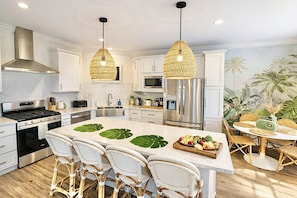 A modern kitchen with a central island, stainless steel appliances, wicker chairs, and a tropical mural.