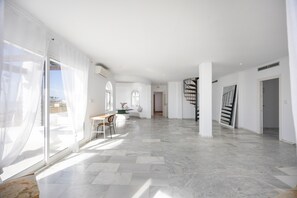 Un interior amplio con piso de mármol, un comedor con mesa y sillas, una escalera y ventanas grandes con cortinas traslúcidas.