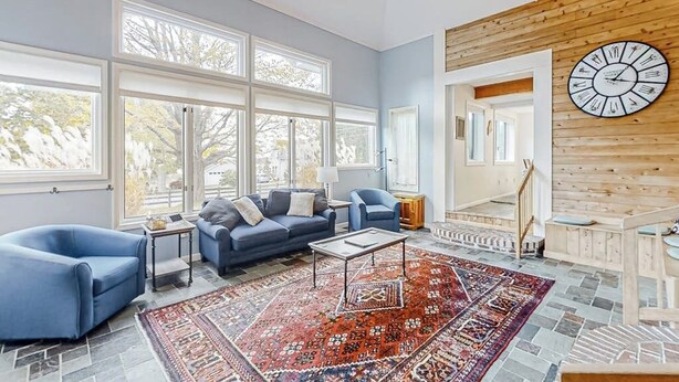 Ein geräumiges Wohnzimmer mit großen Fenstern, einem blauen Sofagarnitur, einem gemusterten Teppich und einer Holzwand mit einer Uhr.