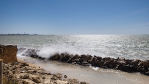 A rocky shoreline with waves crashing against a breakwater, a clear sky, and a distant city skyline. - Isla Verde 3: Sunset Room in a Tranquil Villa in Cadiz (Copina)
