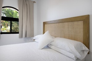 Una cama con sábanas blancas, cabecera de madera y ventana con vistas al follaje.