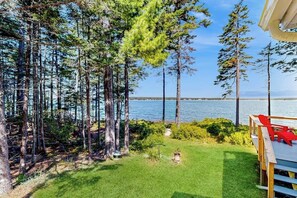 A deck with red chairs overlooking a lake with tall trees. - Dog-friendly, waterfront home on St. George River with shore access, great views (St. George)