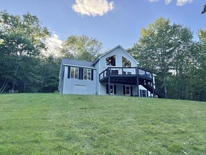 A two-story house with a large deck, a garage, and a well-maintained lawn. - Riverfront home w/ deck, amazing views & firepit - near the best of Maine (Stockton Springs)