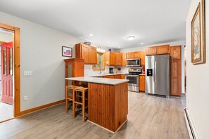 A modern kitchen with wooden cabinets, a stainless steel refrigerator, and a built-in island with stools. - Riverfront home w/ deck, amazing views & firepit - near the best of Maine (Stockton Springs)