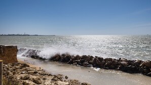Una costa rocosa con olas rompiendo contra un rompeolas, un cielo despejado y un horizonte urbano lejano.