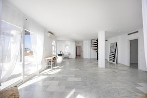 Un interior amplio con piso de mármol, un comedor con mesa y sillas, una escalera y ventanas grandes con cortinas traslúcidas.