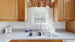 A kitchen with wooden cabinets, a double sink, a window with a floral valance, a toaster, and a dish soap bottle. - Perfect Cottage Near Downtown & Glen Mary Park w/ Full Kitchen, Free WiFi, More (Bar Harbor)
