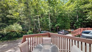 A deck with a table and chairs, surrounded by dense forest and parked cars. - Perfect Cottage Near Downtown & Glen Mary Park w/ Full Kitchen, Free WiFi, More (Bar Harbor)