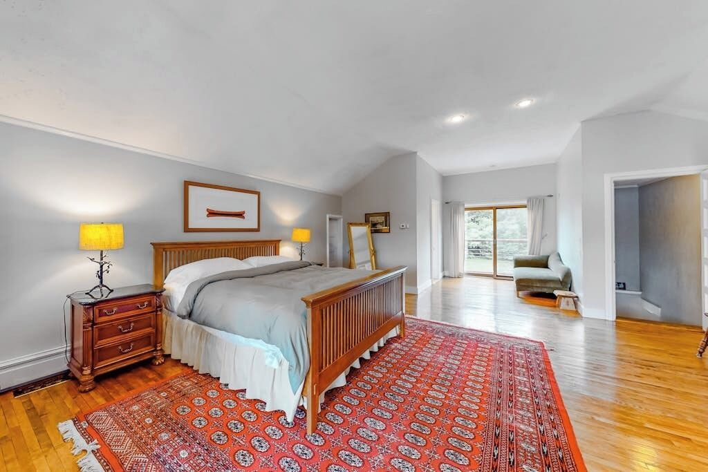 A bedroom with a bed, bedside tables, a red patterned rug, a wooden headboard, and a view to a balcony.