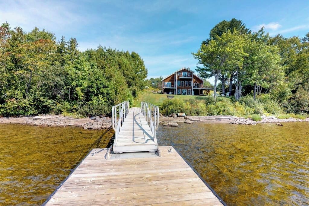 Un muelle de madera que lleva a una casa rodeada de árboles frondosos y un río tranquilo.