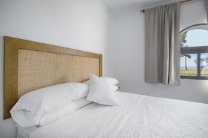 Een net opgemaakt bed met witte lakens en een houten hoofdbord, een raam met uitzicht op het strand, en een gordijn voor het raam.