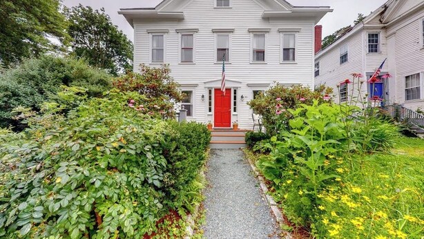A white two-story house with a red door, surrounded by lush greenery and a gravel path leading to the entrance.