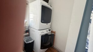 A compact laundry area with stacked washing machines and a dryer, a trash bin, and a wall-mounted ironing board. - Downtown Studio w/ Full Kitchen & Shared Porch - Walk Everywhere in Town (Castine)