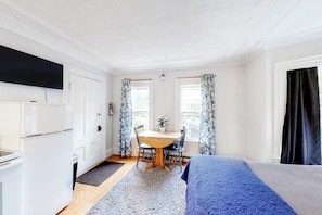 A room with a bed, a dining table with chairs, a TV mounted on the wall, and a window with curtains. - Downtown Studio w/ Full Kitchen & Shared Porch - Walk Everywhere in Town (Castine)