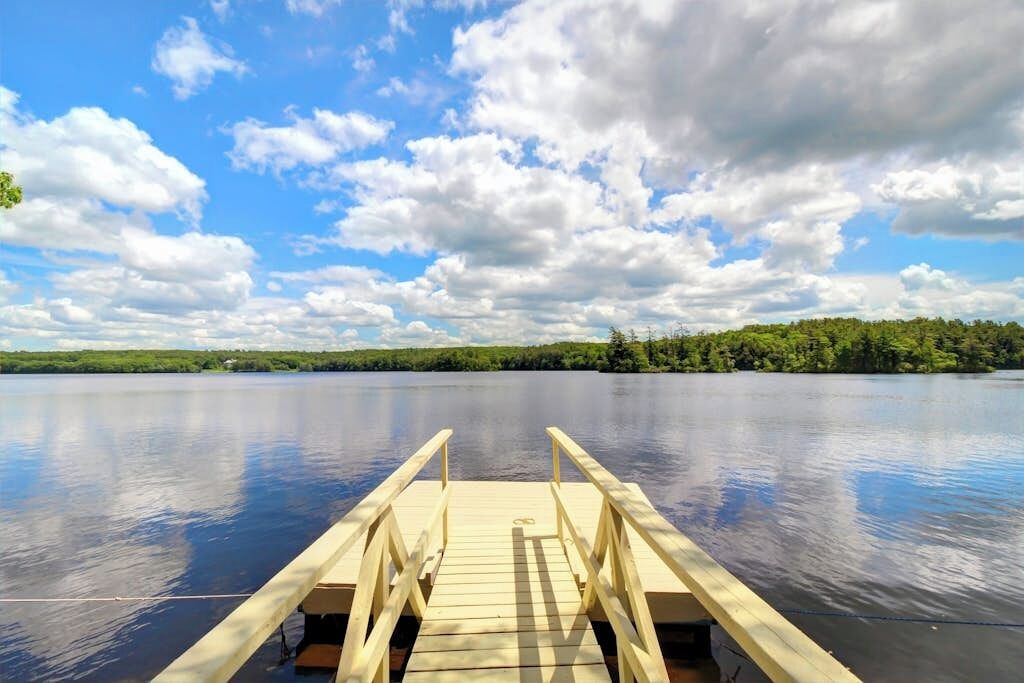A wooden dock extending into a calm lake with a forested shoreline under a partly cloudy sky.