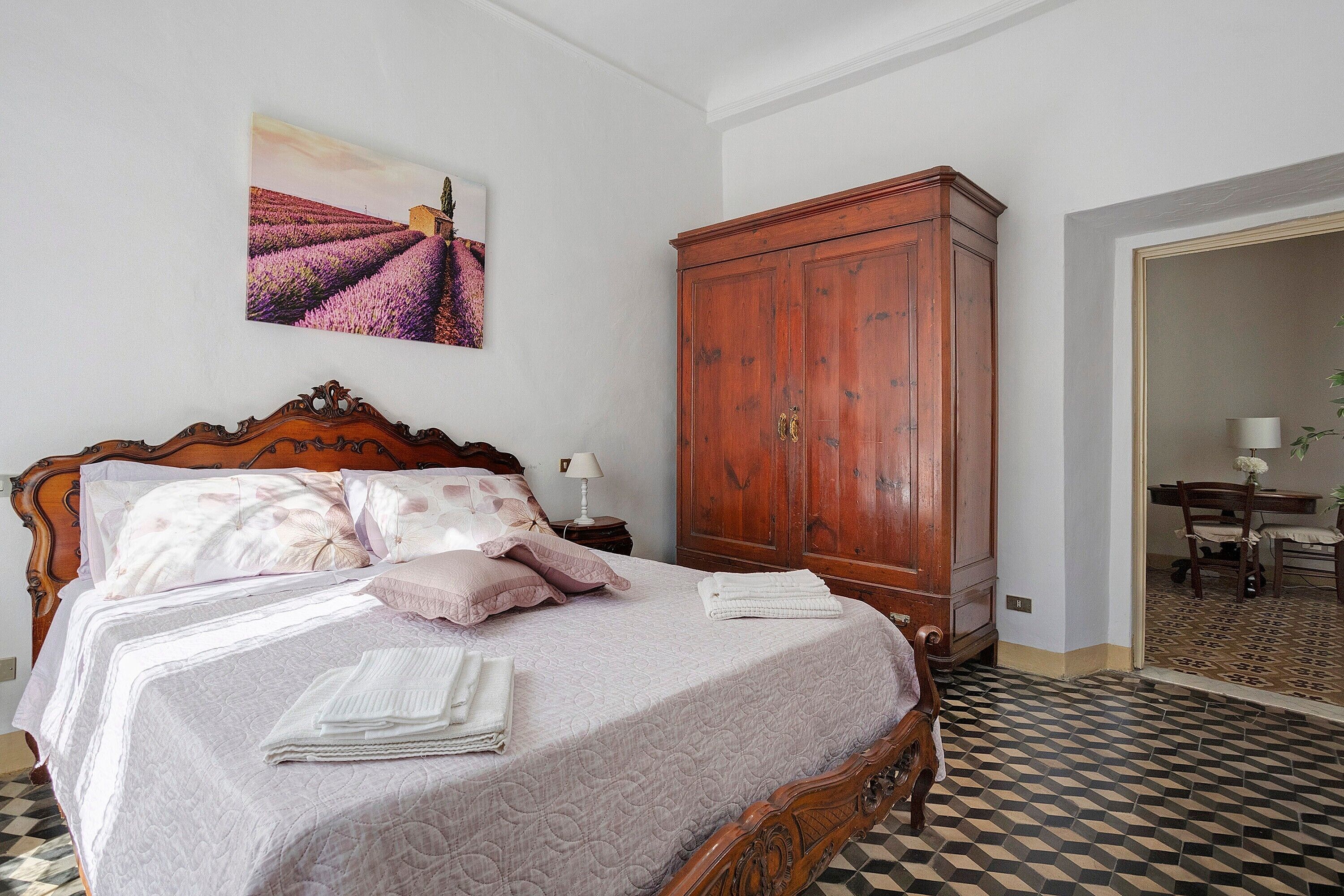 Un dormitorio con cama, un armario de madera, un cuadro en la pared y una zona de comedor visible a través de una puerta abierta.