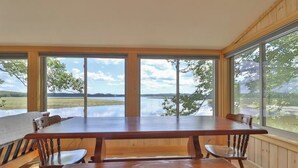 A wooden dining table with benches, large windows, and a view of a lake and hills. - Luxe Bayfront Cabin w/ Stunning Views & Shared Dock- Ideal for Hunting & Fishing (Bowdoinham)