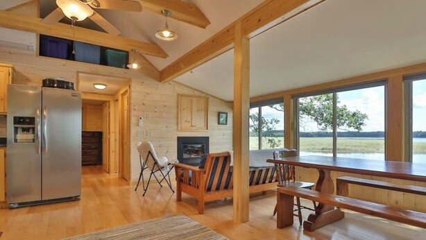 A spacious living area with a wooden floor, a large window overlooking a lake, a fireplace, and a dining table. - Luxe Bayfront Cabin w/ Stunning Views & Shared Dock- Ideal for Hunting & Fishing (Bowdoinham)