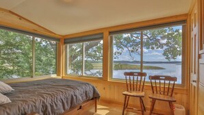 A bedroom with a bed, two chairs, a table, and large windows with a view of a lake and trees. - Luxe Bayfront Cabin w/ Stunning Views & Shared Dock- Ideal for Hunting & Fishing (Bowdoinham)