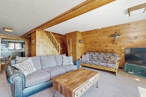 A cozy cabin interior with a sofa, coffee table, wooden walls, a staircase, and a kitchen area. - Comfy Lakefront Cabin with Gorgeous Views, Private Dock & Pebble Beach (Lily Bay Township)