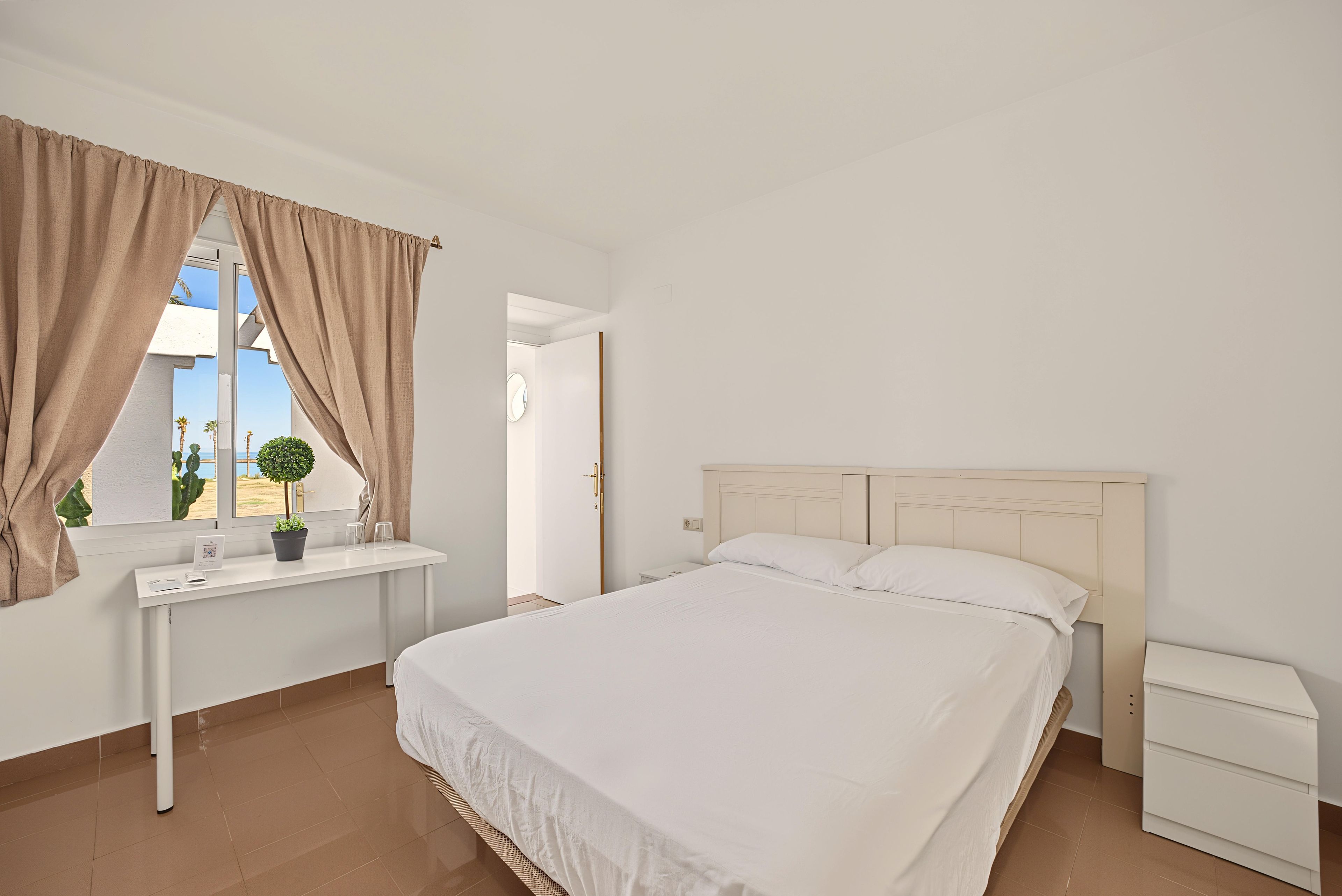 Een slaapkamer met een bed, nachtkastje, raam met gordijnen, een kleine potplant en uitzicht op het landschap.