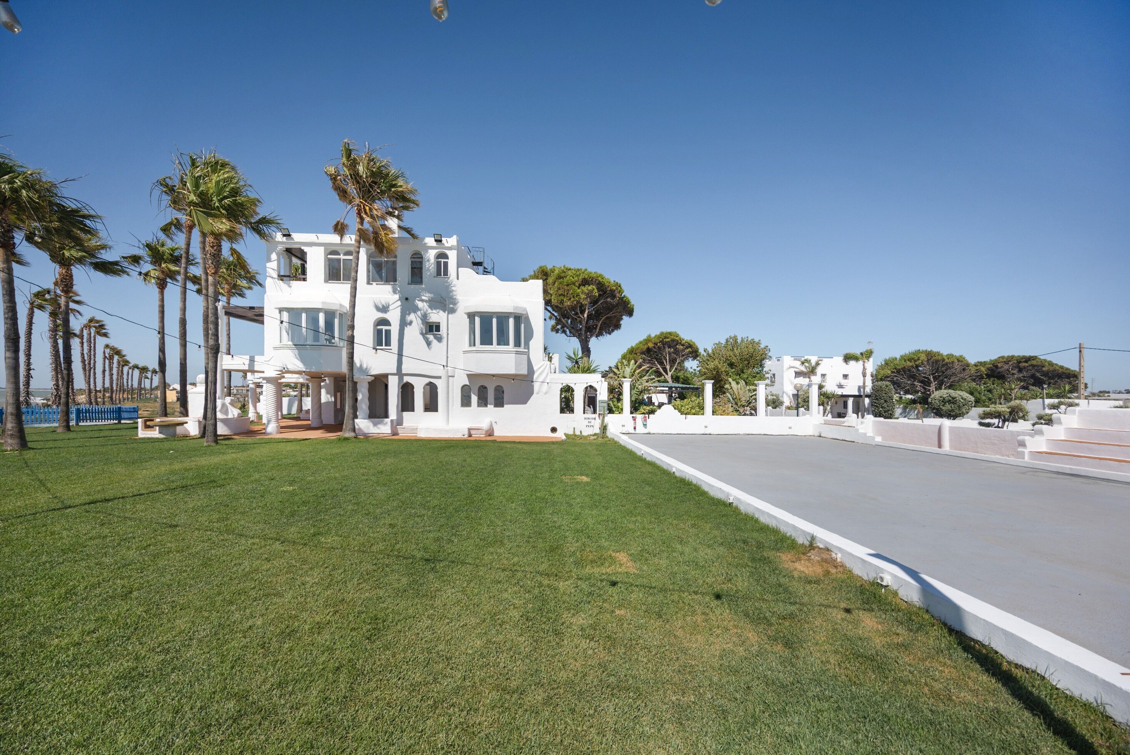 A white multi-story building with balconies, surrounded by a well-maintained lawn and palm trees.