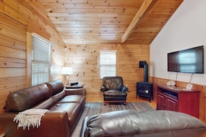 Ein Blockhaus-Interieur mit Ledersofa, Ohrensessel, Hocker, Kamin, Fernseher und Holzwänden.