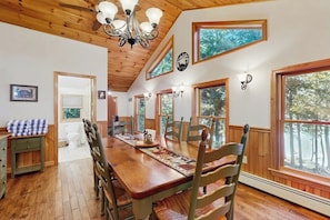 A dining area with a wooden table, chairs, a chandelier, and large windows with a view of trees. - Lakefront cabin close to town with dock, firepit - dogs welcome (Beaver Cove)