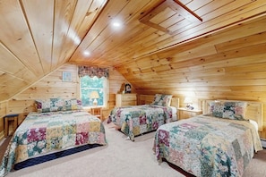 A cozy attic bedroom with wooden walls and sloped ceiling, featuring three beds with floral bedding and a small table with a lamp. - Lakefront cabin close to town with dock, firepit - dogs welcome (Beaver Cove)
