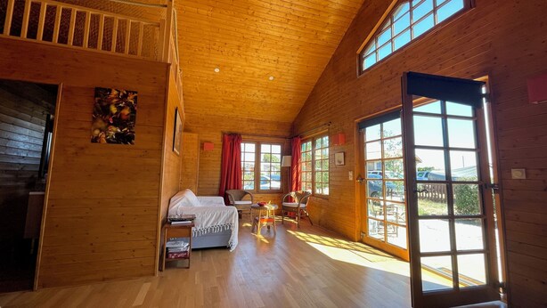 A spacious room with wooden floors, a bed, a chair, a table, and large windows. - Wooden beach house for vacations - Le Verdon-sur-Mer (33) (Le Verdon-sur-Mer)