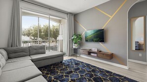 Una sala moderna con un sofá seccional, un televisor montado en la pared, una mesita auxiliar con una planta y una alfombra estampada.