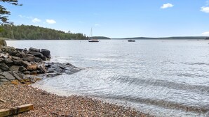 A calm lake with boats, a rocky shoreline, and a forested hill in the background. - Comfortable bayfront home with beautiful views, large deck & on-site tennis (Seal Cove)