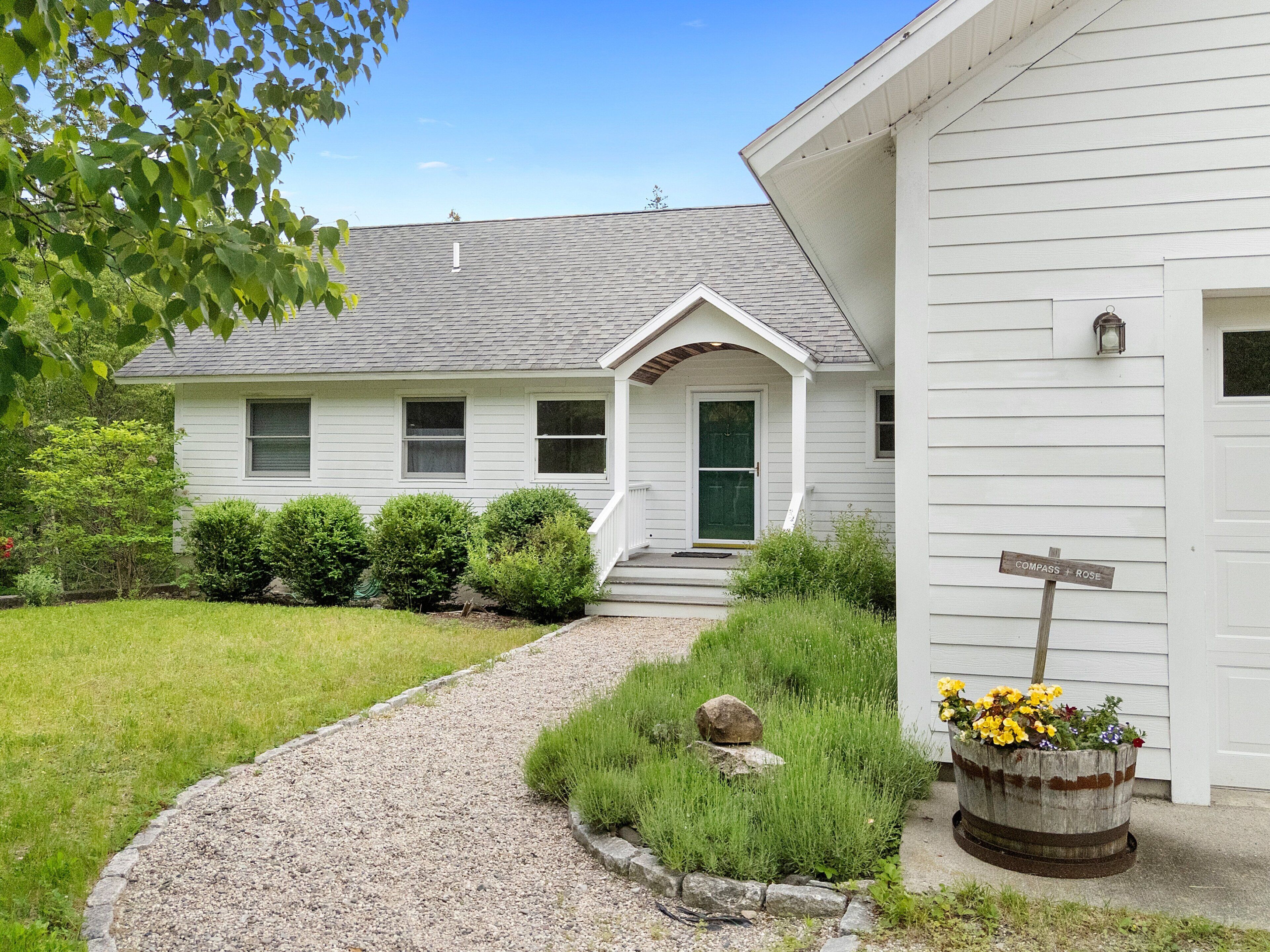 A white house with a gravel path leading to the front door, surrounded by greenery and a flower pot.