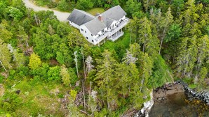 An aerial view of a large white house with a spacious porch and multiple windows, surrounded by dense forest. - Comfortable bayfront home with beautiful views, large deck & on-site tennis (Seal Cove)