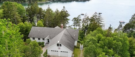 A house with a garage, surrounded by dense forest, near a large body of water.