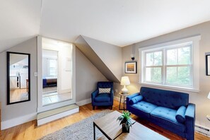 Un salon avec un canapé bleu, un fauteuil bleu, une table basse ornée d’une plante, une lampe et une grande fenêtre donnant sur un beau paysage.